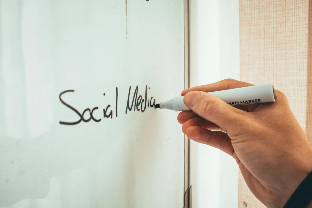 Close-up of a hand using a black marker to write "Social Media" on a glass whiteboard.